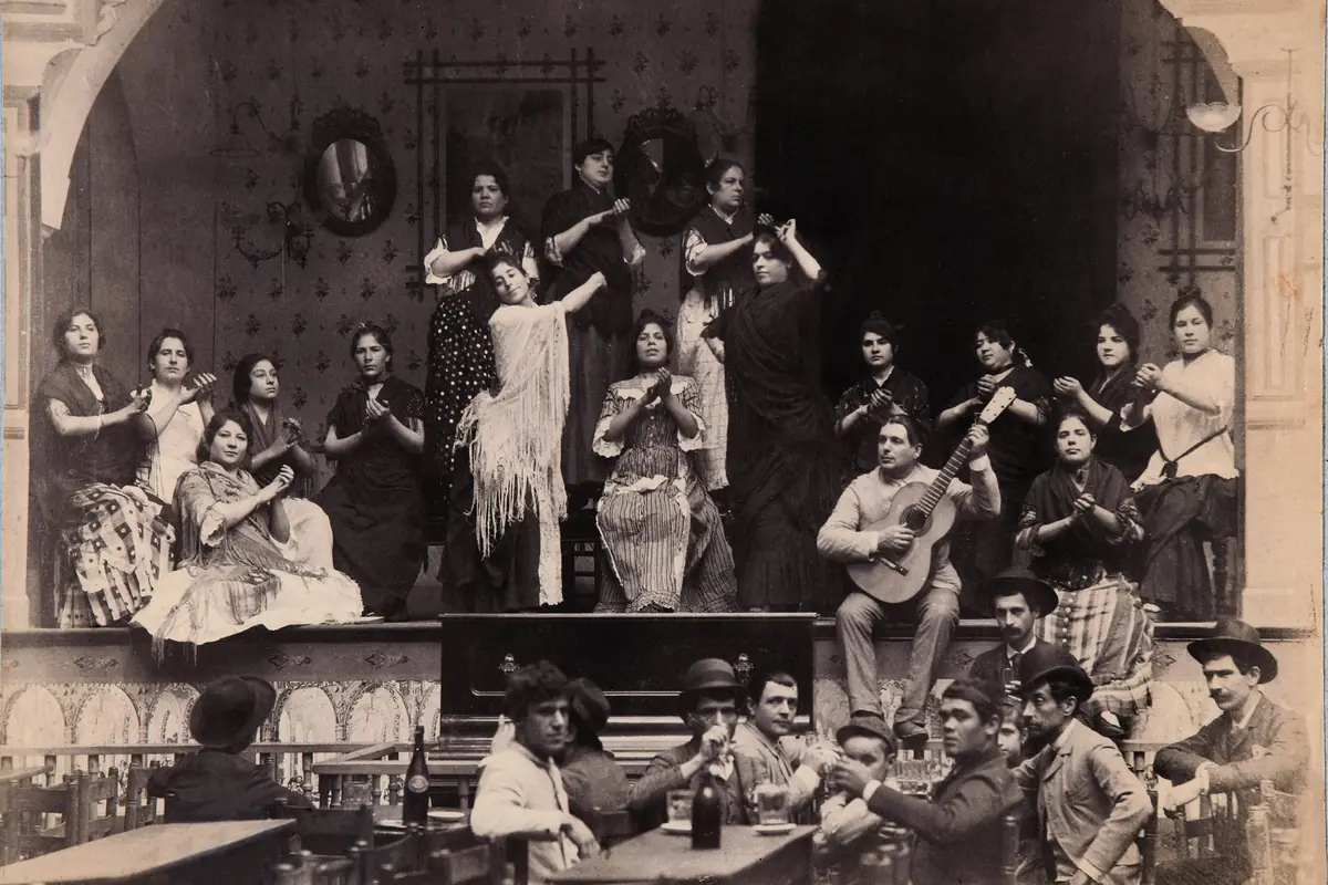 Sepiová fotografie pódia café cantante v Seville, se ženami v šálách, které tleskají a tančí, sedícím kytaristou a muži v kloboucích popíjejícími u stolů vpředu