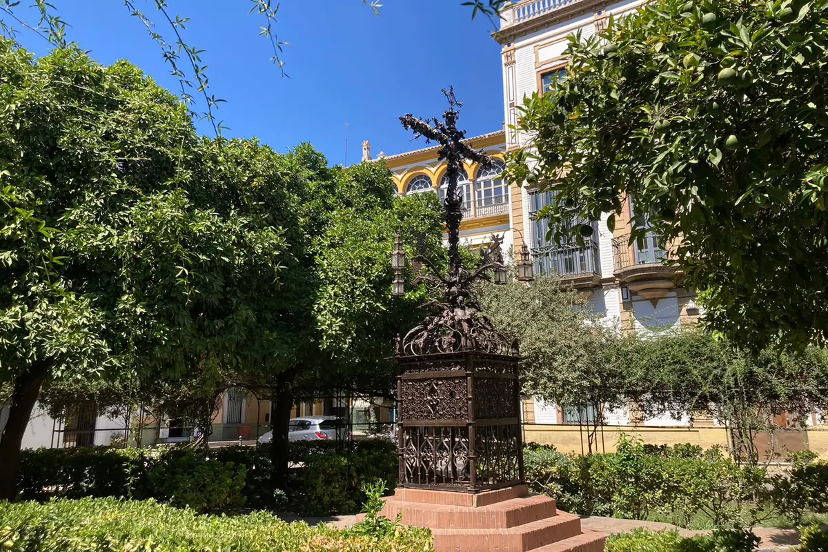 Vysoký kovaný kříž Cruz de la Cerrajería na cihlovém podstavci na náměstí Plaza de Santa Cruz, orámovaný pomerančovníky a bílými a okrovými domy pod modrým nebem